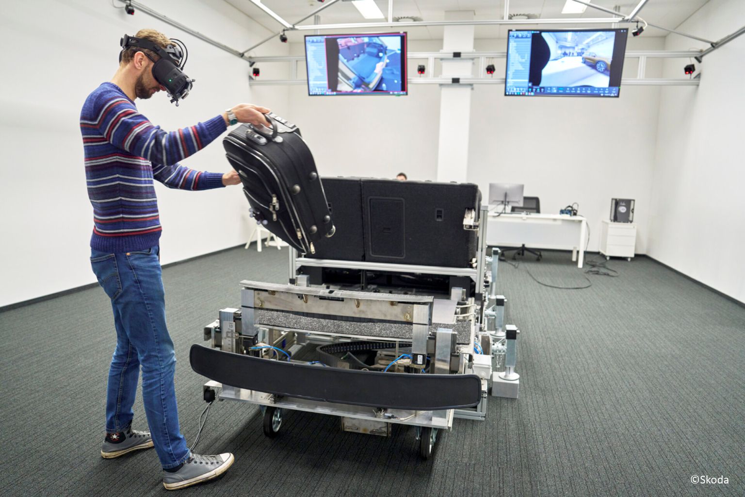 VR-Ergonomietest bei Škoda: Person mit Headset hebt einen Koffer in ein Kofferraum-Mock-up; Decken-Tracking und Monitore im Raum.